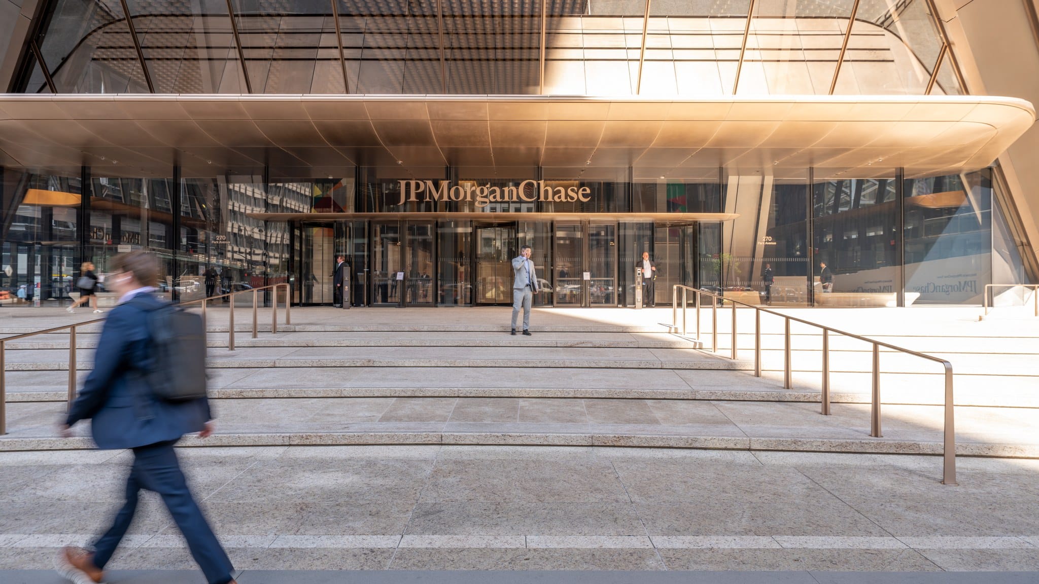 The glass-fronted entrance of JPMorgan Chase's headquarters at 270 Park Avenue in New York, with wide stone steps and a pedestrian walking past.