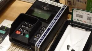 A card payment terminal with keypad and contactless reader beside a QR code scanner on a shop counter in Japan.