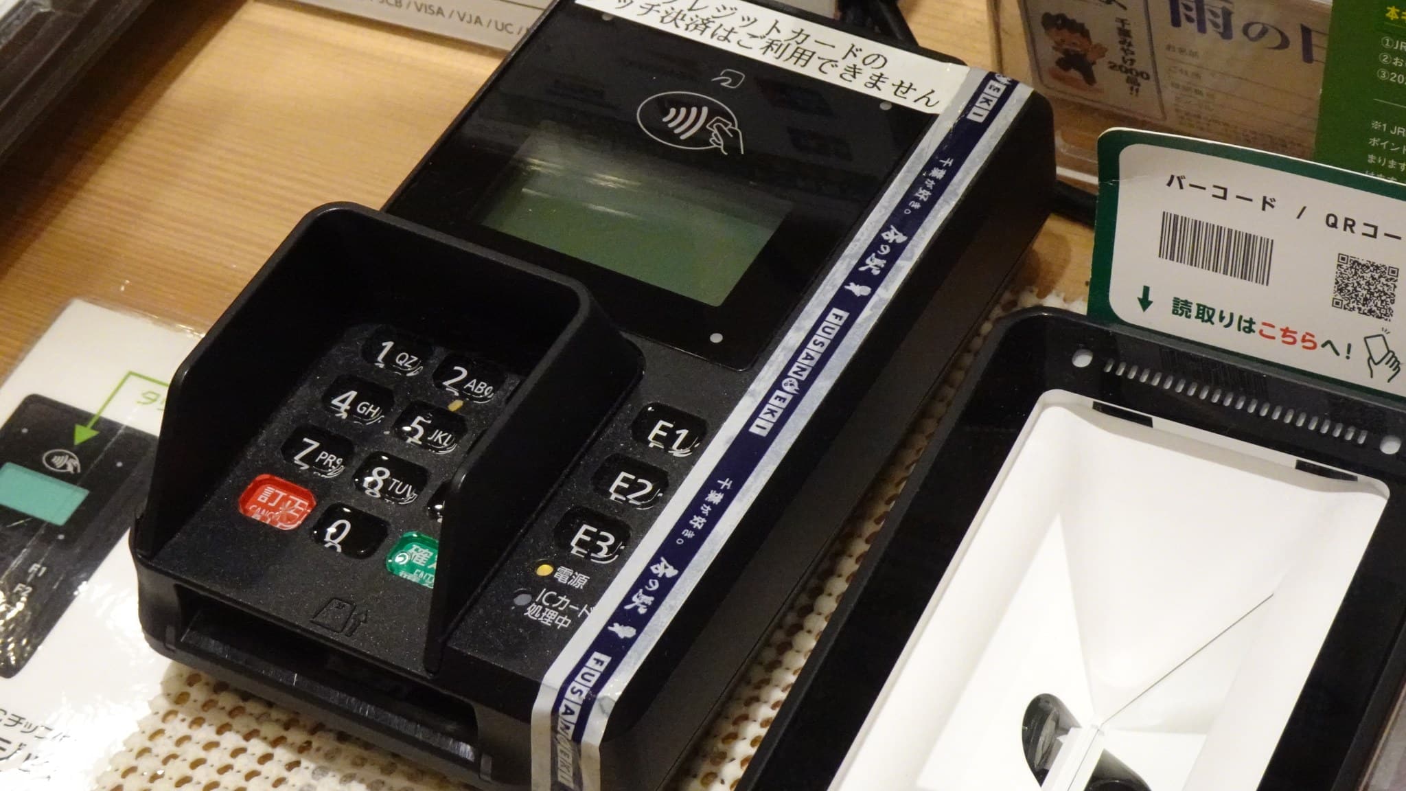 A card payment terminal with keypad and contactless reader beside a QR code scanner on a shop counter in Japan.