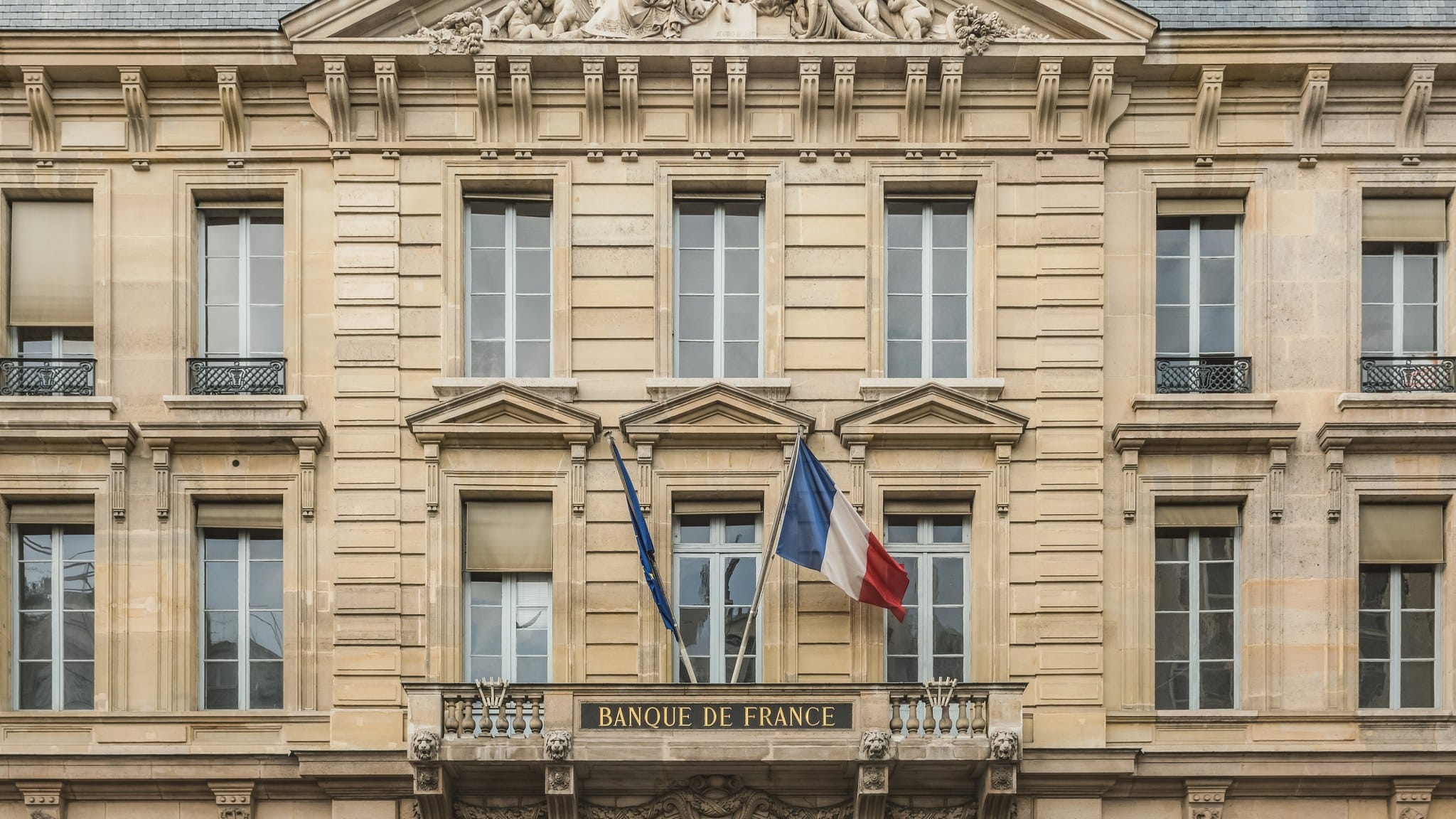 Stone facade of the Hotel de Toulouse in Paris, headquarters of the Banque de France, with the French and European Union flags above a balcony bearing the words Banque de France.