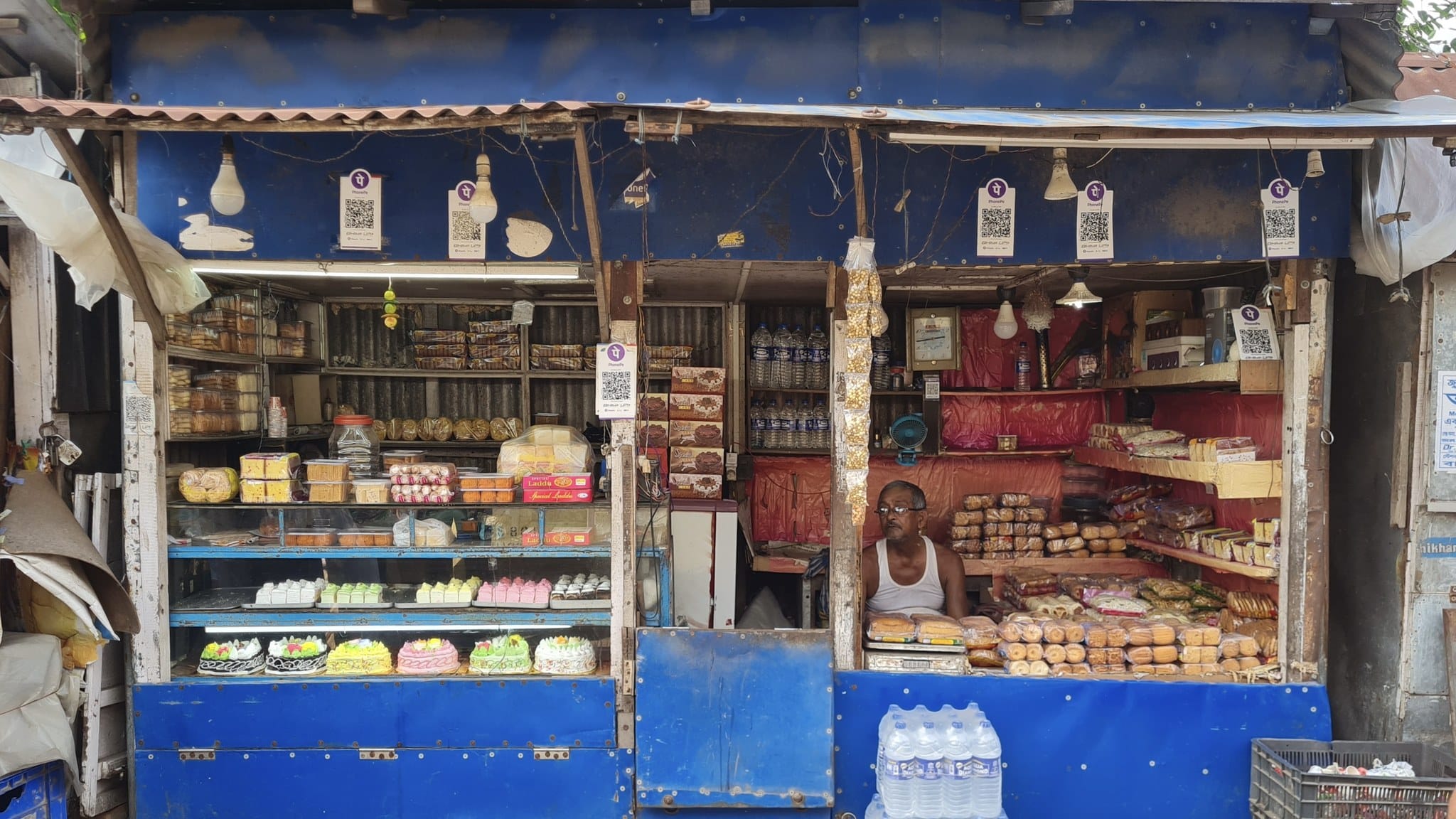 A small sweet shop in North 24 Parganas, West Bengal, with several QR code payment placards hanging along its front and a shopkeeper behind the counter.